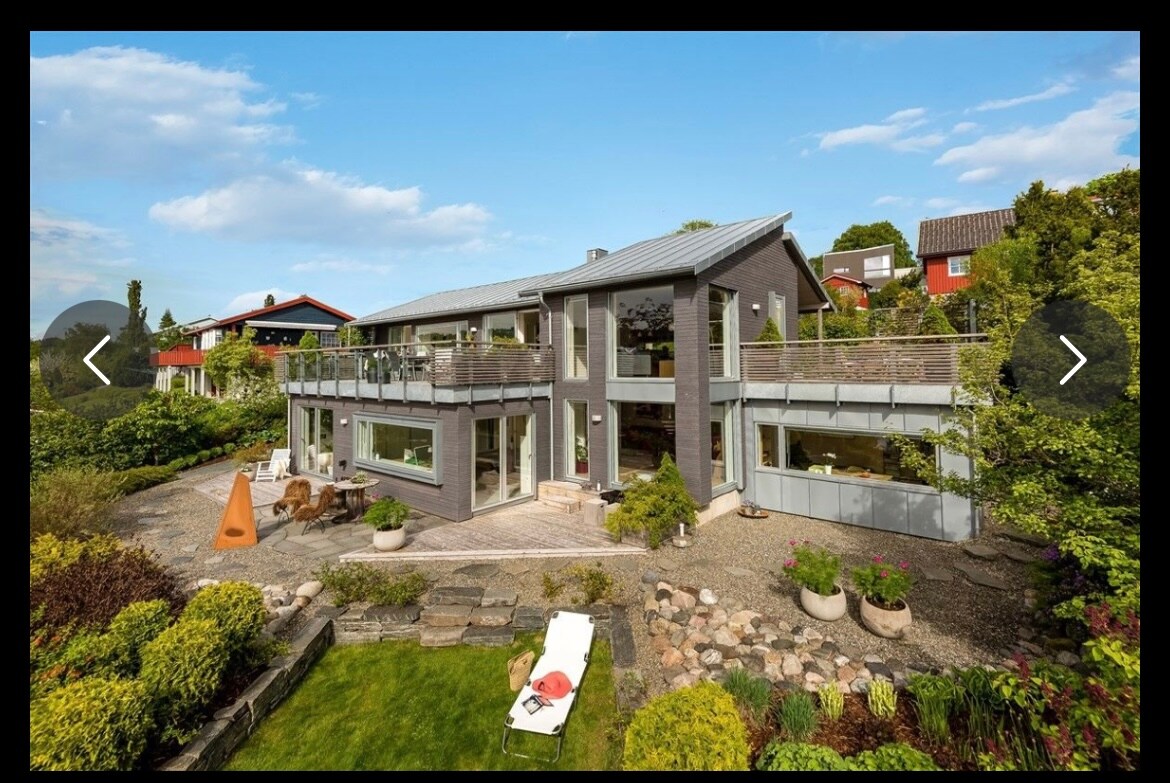 The exterior of the home features a modern design with large glass windows and a spacious terrace. Surrounding greenery includes flowering plants and well-maintained pathways. A lounge chair is positioned on the lawn, inviting relaxation in the tranquil outdoor space.