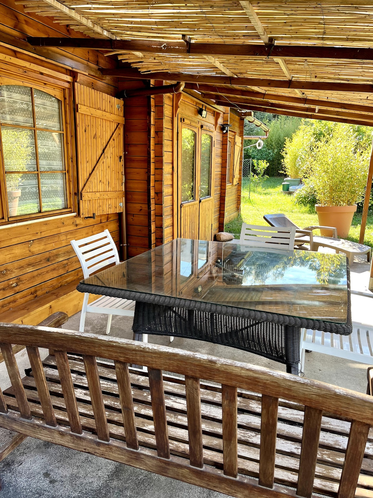 An outdoor seating area features a glass-topped table surrounded by white chairs and a wooden bench. The rustic wooden walls create a warm backdrop, with greenery visible in the distance, enhancing the natural ambiance of the space.