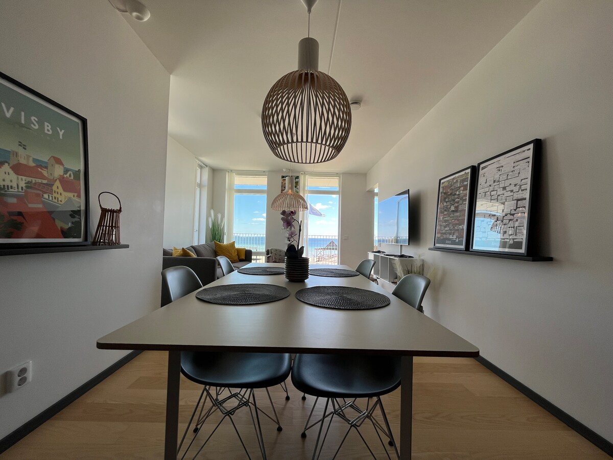 A dining area is highlighted by a modern table set for four, featuring round placemats and a stylish pendant light overhead. Large windows provide a view of the outside, while art pieces are displayed on the walls, enhancing the contemporary decor.