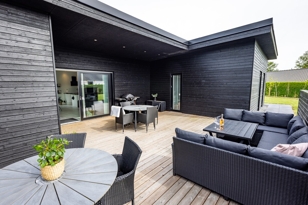 A spacious outdoor terrace features a blend of lounge and dining furniture, providing an inviting area for relaxation. The terrace is surrounded by greenery, with an open layout and large glass doors leading into the interior. Natural wood tones enhance the overall warm atmosphere.