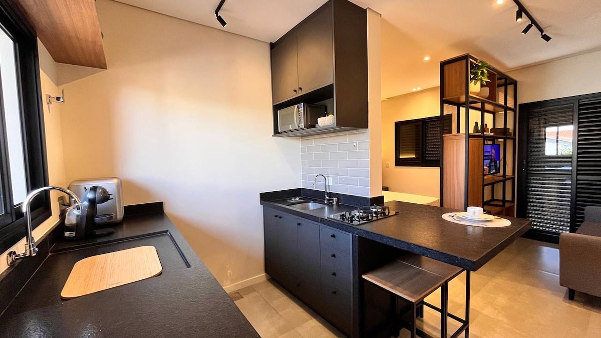 A modern kitchen is featured, equipped with dark cabinetry and a sleek black countertop. Stainless steel appliances, including a microwave and a stovetop, are visible. A small table is present, positioned near a window letting in natural light.