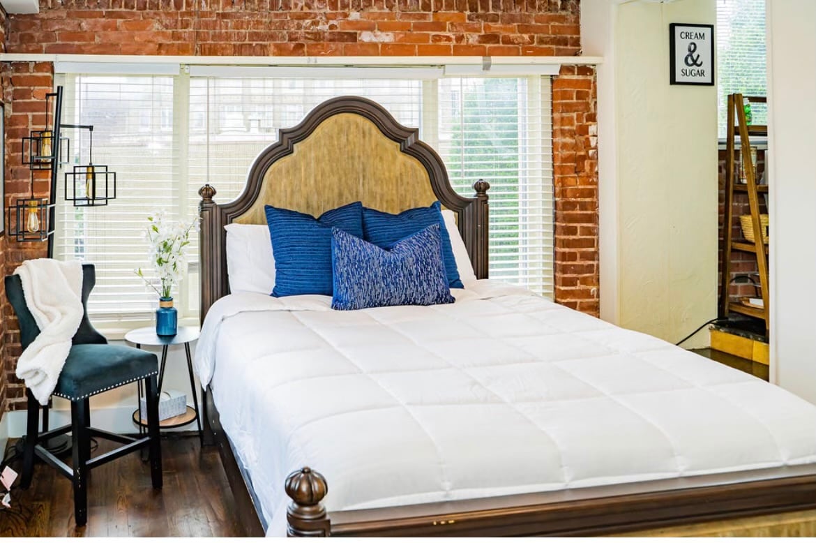 A spacious bed is centered against a backdrop of exposed brick walls, complemented by large windows allowing natural light to enter. Decorative throw pillows in shades of blue are arranged on the bed. A small side table holds a vase with flowers, and a chair is positioned nearby.