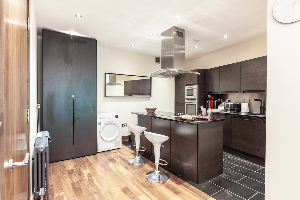 A modern kitchen space showcases dark cabinetry and sleek appliances, including a built-in oven and microwave. An island bar with two stools offers a casual dining area. A washer is positioned to the side, and large windows provide ample natural light, brightening the room.