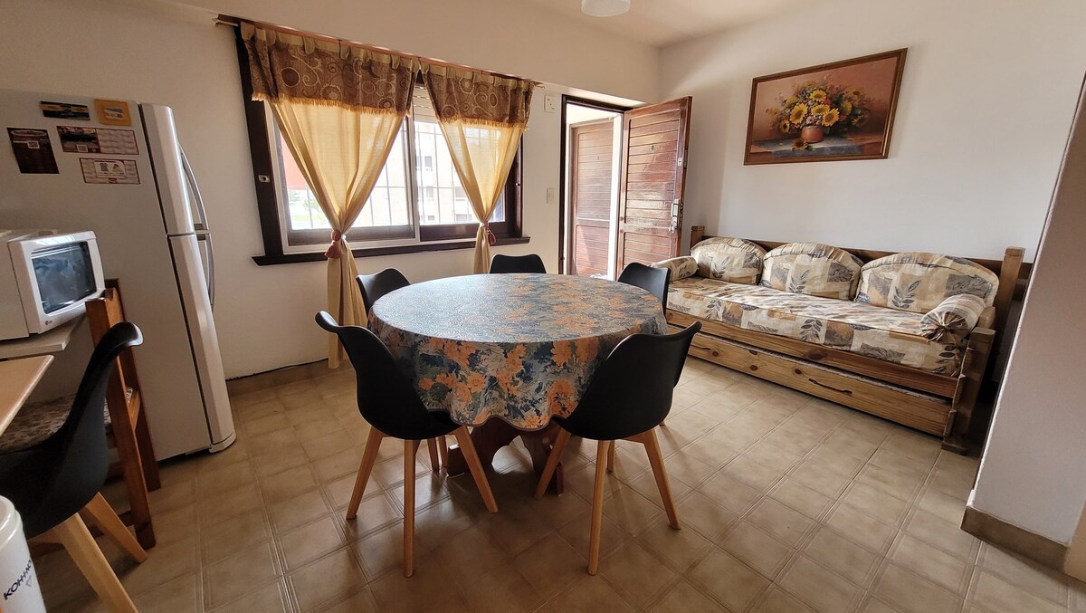 A dining area is presented, featuring a round table surrounded by four black chairs. A beige sofa with patterned fabric is placed against the wall. Large windows are draped with light curtains, allowing natural light to brighten the space.