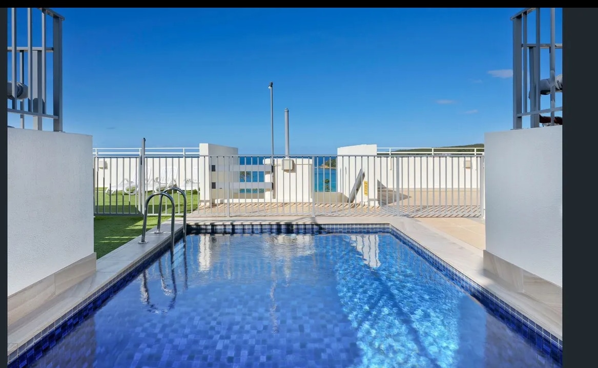 A sparkling pool with a deep blue tiled surface is highlighted under a clear blue sky. Surrounding area includes lounge chairs and a fenced terrace, providing clear views and ample space for relaxation.