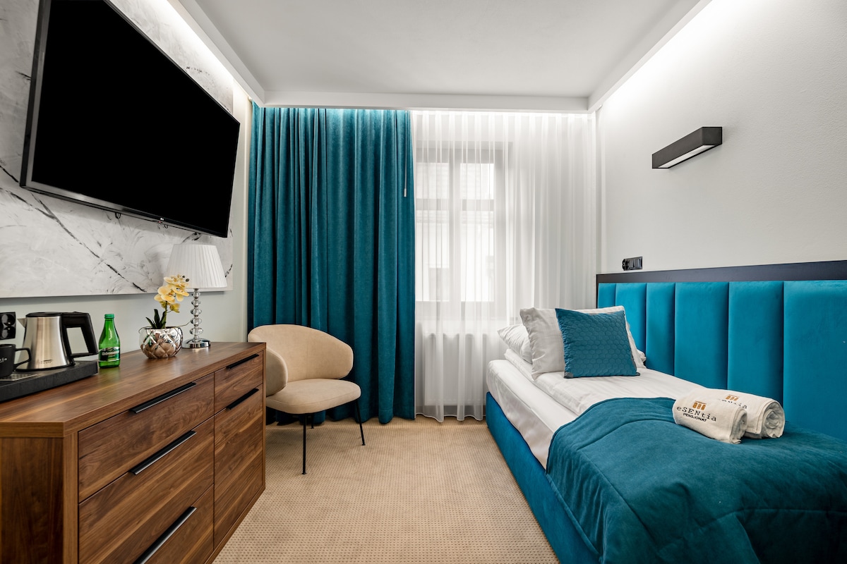 A modern single room features a spacious bed, accented by teal bedding and cushions. A light-colored armchair sits near the window, flanked by sheer curtains. A sleek wooden dresser displays a coffee and tea setup, while a large wall-mounted television is positioned for viewing.