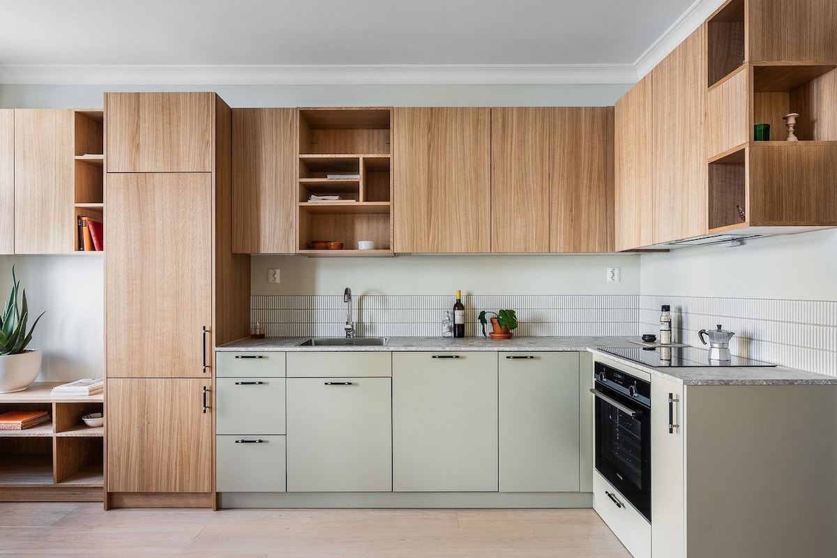 A modern kitchen displays sleek cabinetry in light and wooden finishes. Counter space is complemented by minimalist appliances, including an oven and a sink. Shelves house various items, while a small plant adds a touch of greenery. The overall layout encourages functionality and convenience.