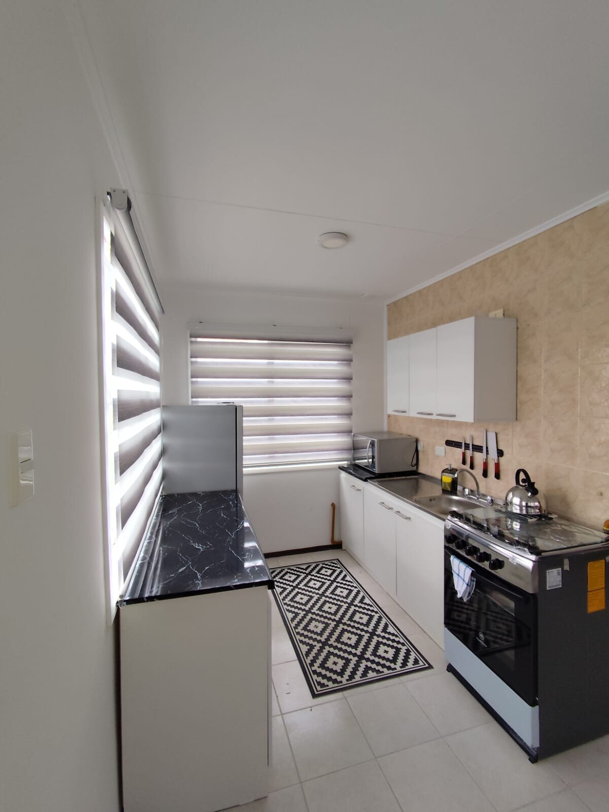 A compact kitchen is equipped with modern appliances, featuring a sleek countertop and cabinetry. Natural light filters through patterned blinds, illuminating the space. A patterned rug adds warmth to the floor, while utensils are organized neatly on the wall.