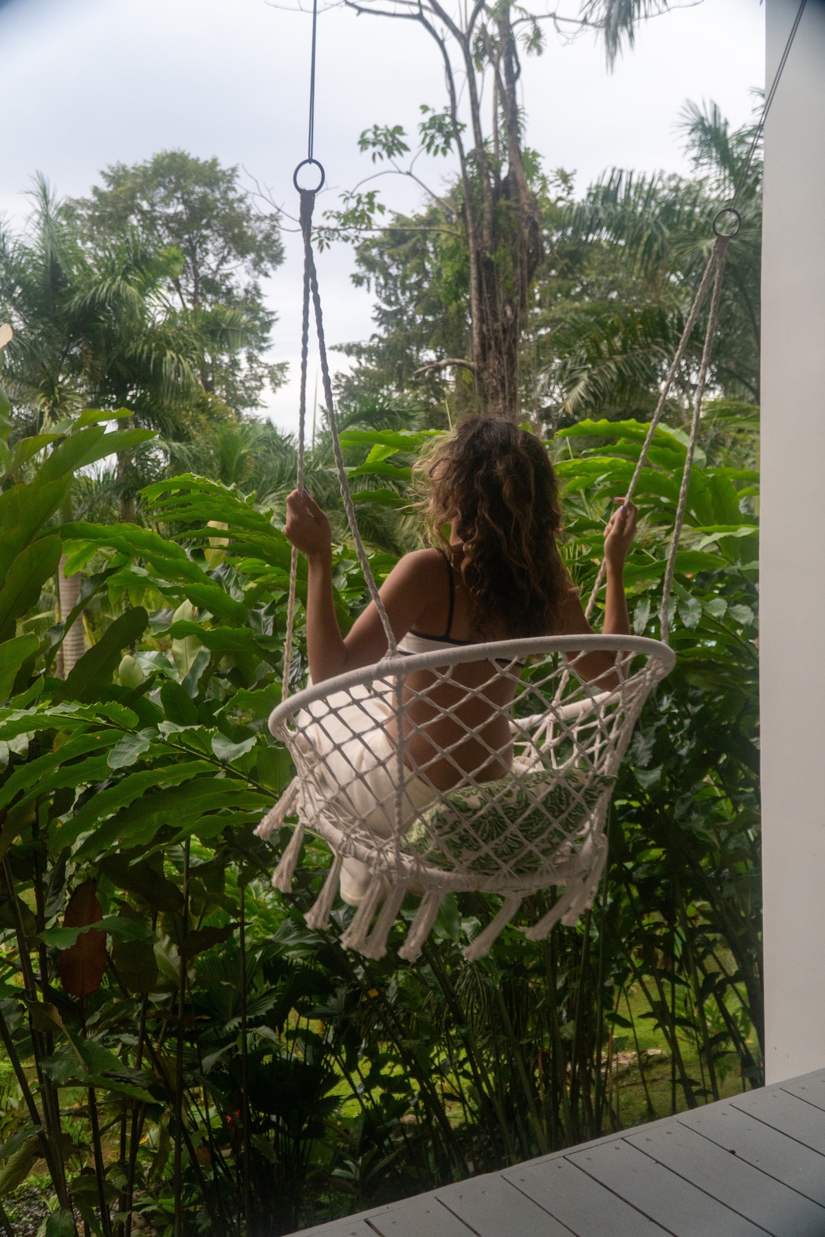 A white macrame swing hangs from a porch, offering a serene view of verdant jungle foliage. Lush green plants surround the swing, creating a peaceful and immersive atmosphere in the outdoors.