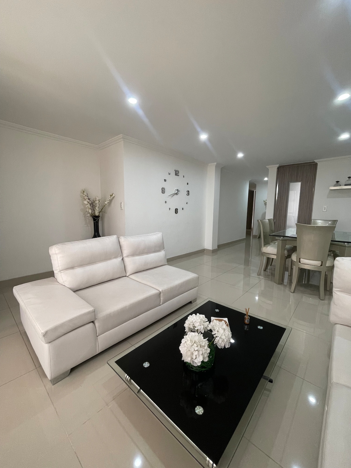 A modern living area is presented with white leather sofas arranged around a dark glass coffee table. A decorative bouquet sits on the table, accompanied by a wall clock above. Neutral-toned walls and glossy tile floors contribute to a clean and open ambiance.