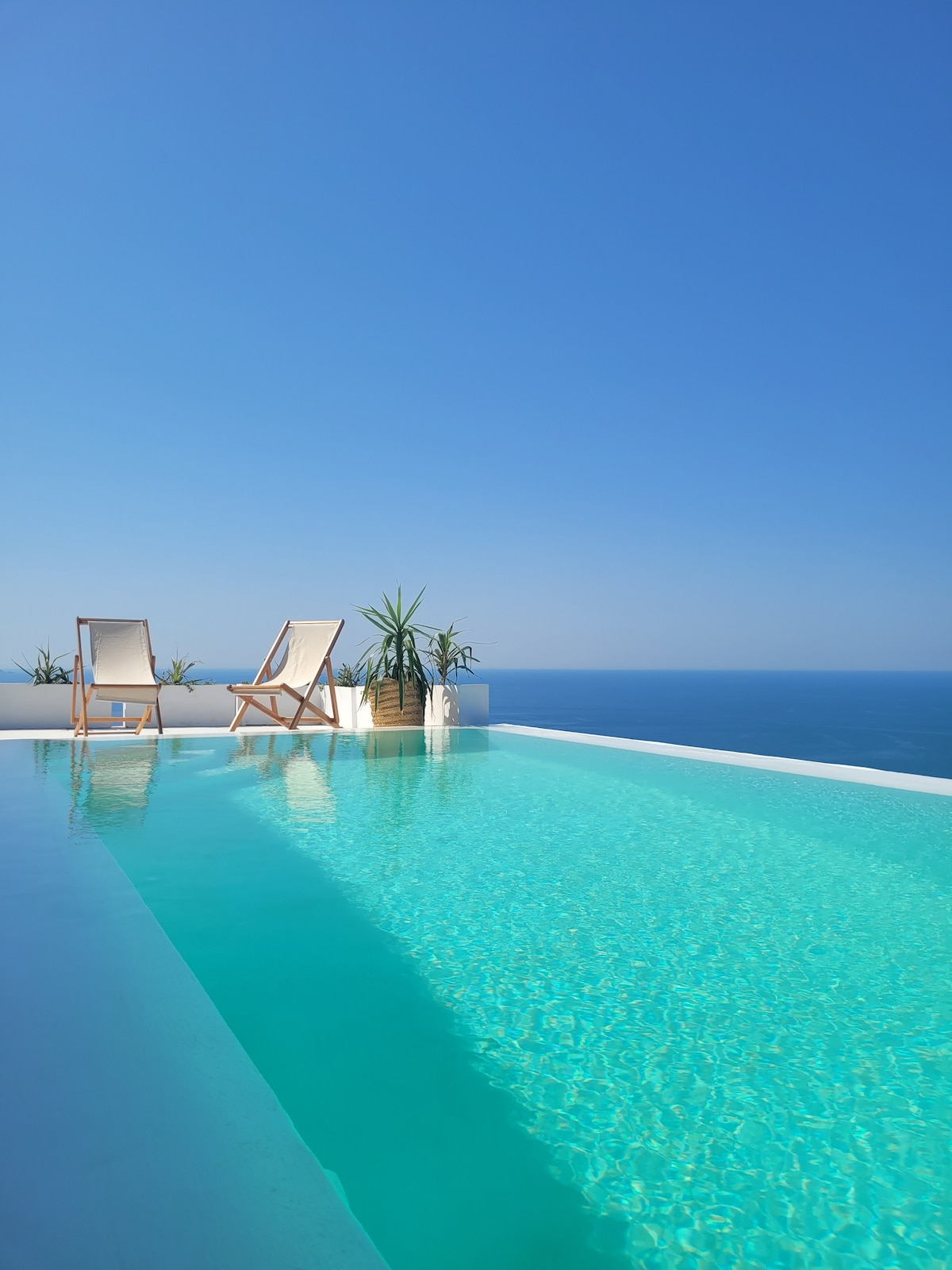 An infinity pool extends towards the horizon, blending with the expansive blue sea. Two wooden lounge chairs rest poolside, accompanied by potted plants. The clear sky and sunlight enhance the tranquil setting.