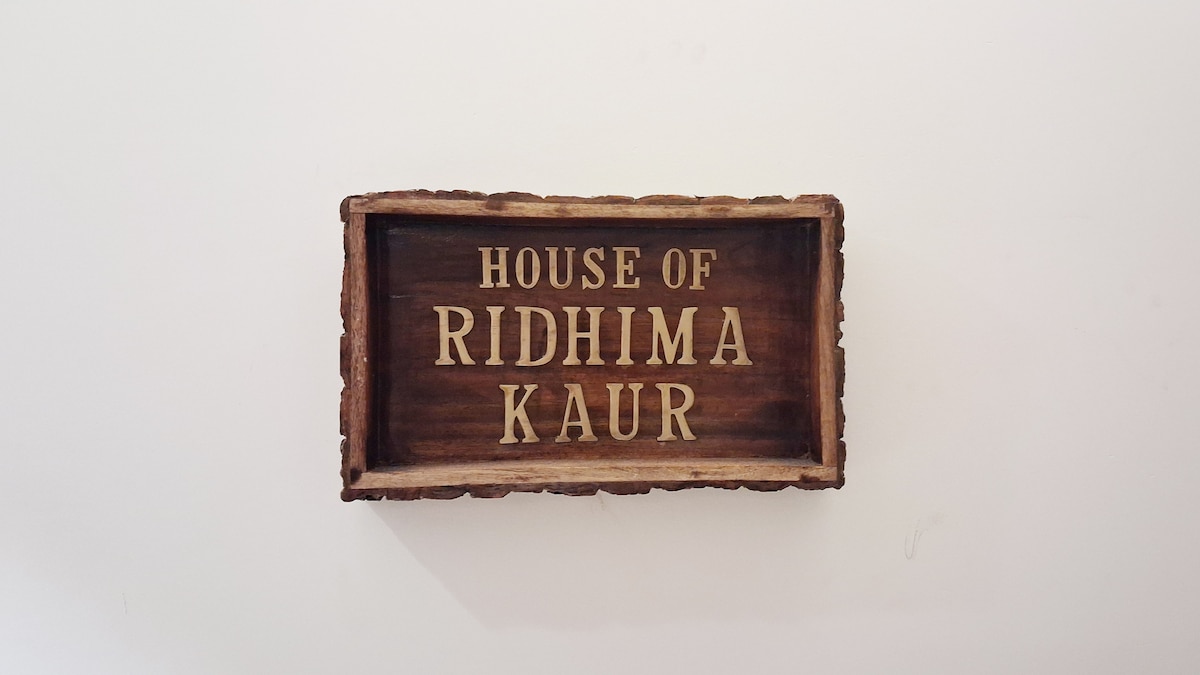 A rustic wooden sign is displayed against a plain white wall, featuring the engraved text 'HOUSE OF RIDHIMA KAUR'. The natural finish of the wood complements the character of the sign, creating a welcoming focal point.