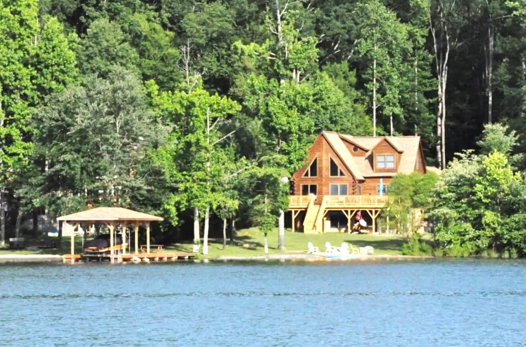A lakeside log home is set against a backdrop of lush greenery, featuring a spacious deck extending from the upper level. A private dock with a covered area is visible in front of the home, offering direct access to the calm waters of the lake.
