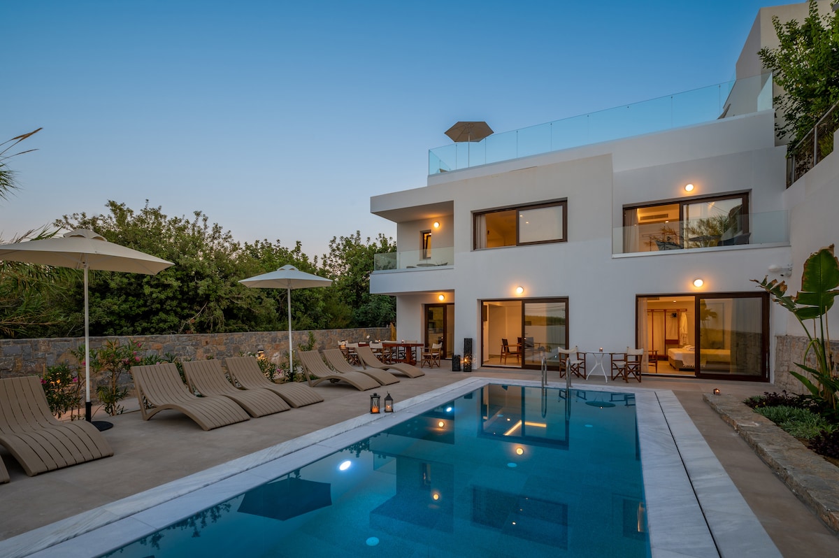 The villa features a private pool surrounded by spacious sun loungers and umbrellas. Large glass doors lead to living areas, showcasing an inviting outdoor space. Lush greenery borders the areas, adding to the serene atmosphere.