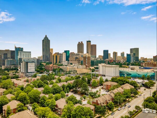 A panoramic view of the Atlanta skyline captures a blend of modern skyscrapers and green spaces. The cityscape features various architectural styles, with lush trees dotting the foreground and a clear blue sky above, showcasing the vibrant urban environment.