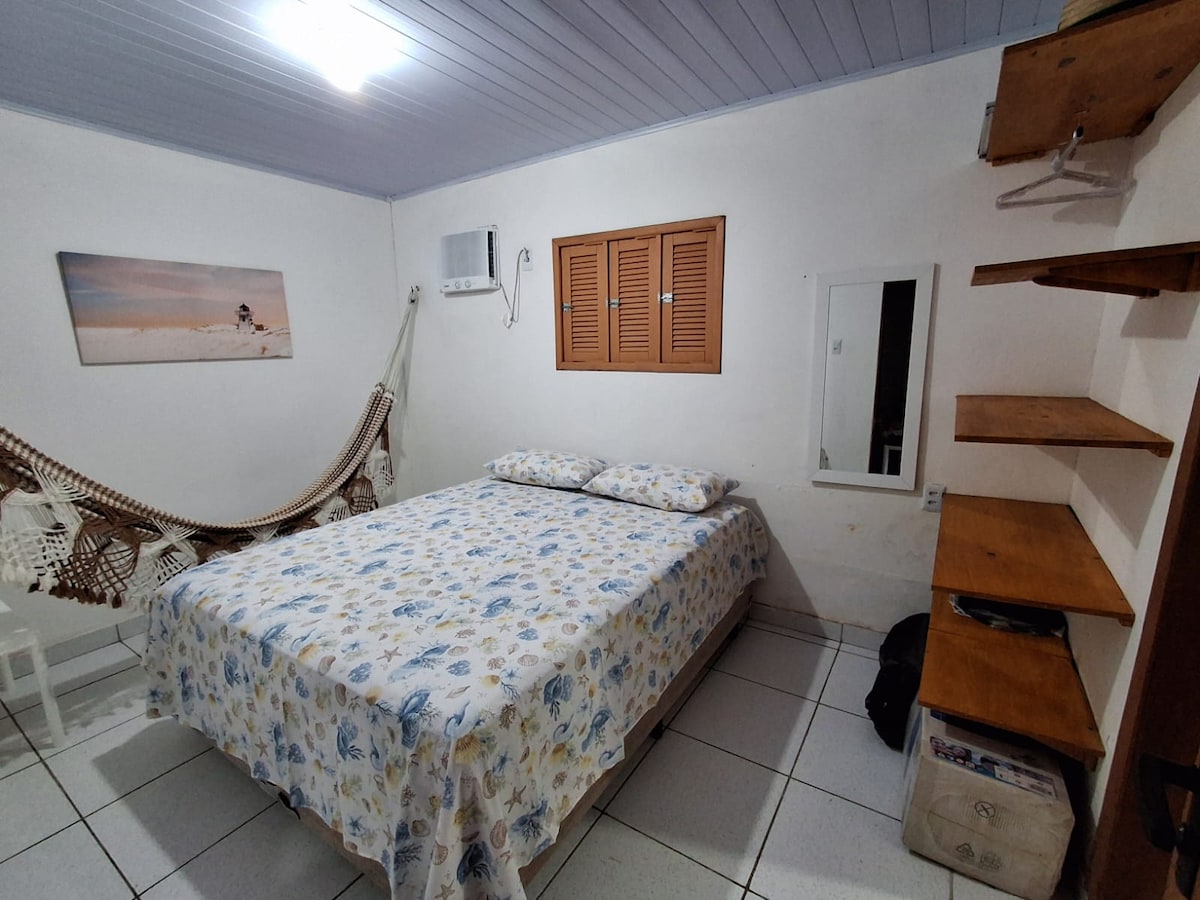 A bright bedroom features a spacious bed dressed in floral bedding. A hammock is hung on one side, while a window with wooden shutters offers natural light. Shelving provides storage, and a mirror contributes to a functional layout.