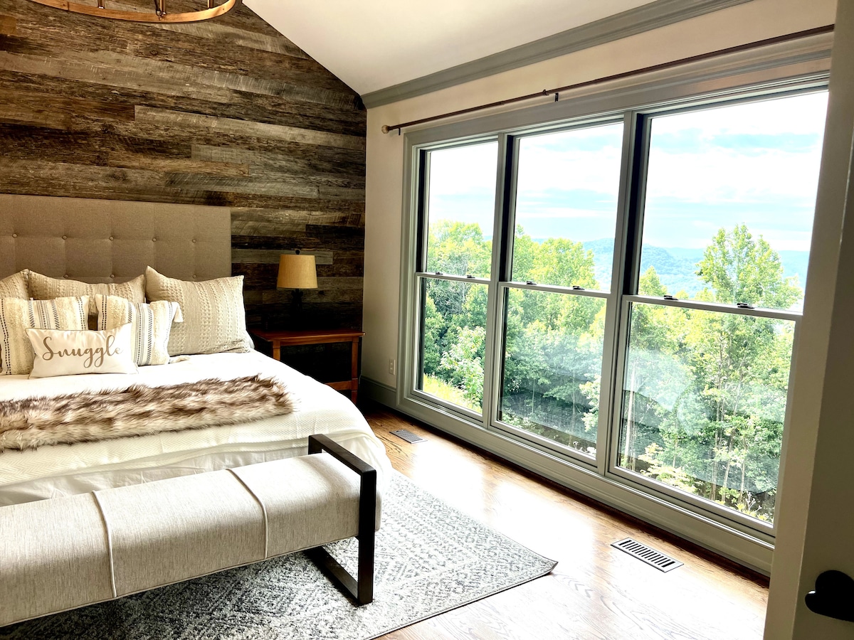 A serene bedroom features a spacious bed adorned with decorative pillows, complemented by a cozy throw. A bench sits at the foot of the bed. Large windows provide a sweeping view of lush greenery and distant mountains, filling the room with natural light.