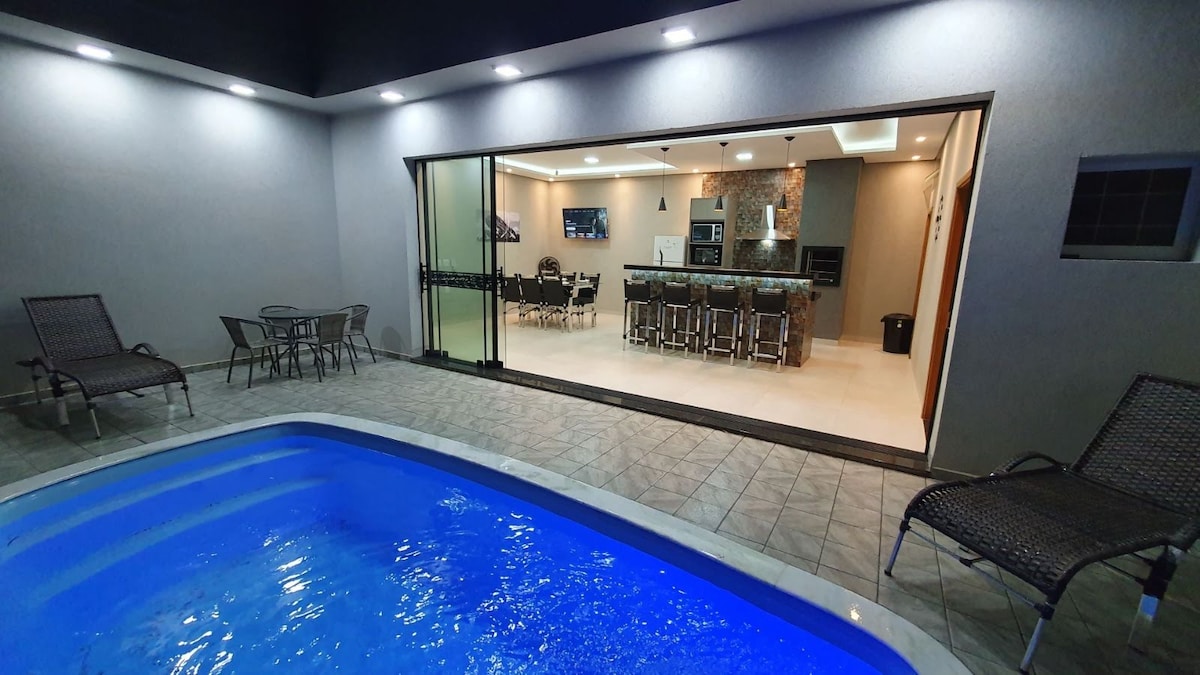 A well-lit outdoor area features a blue swimming pool, complemented by two lounge chairs nearby. The view extends to a spacious indoor area where dining and kitchen spaces are showcased through large sliding glass doors, creating a seamless transition between indoors and outdoors.