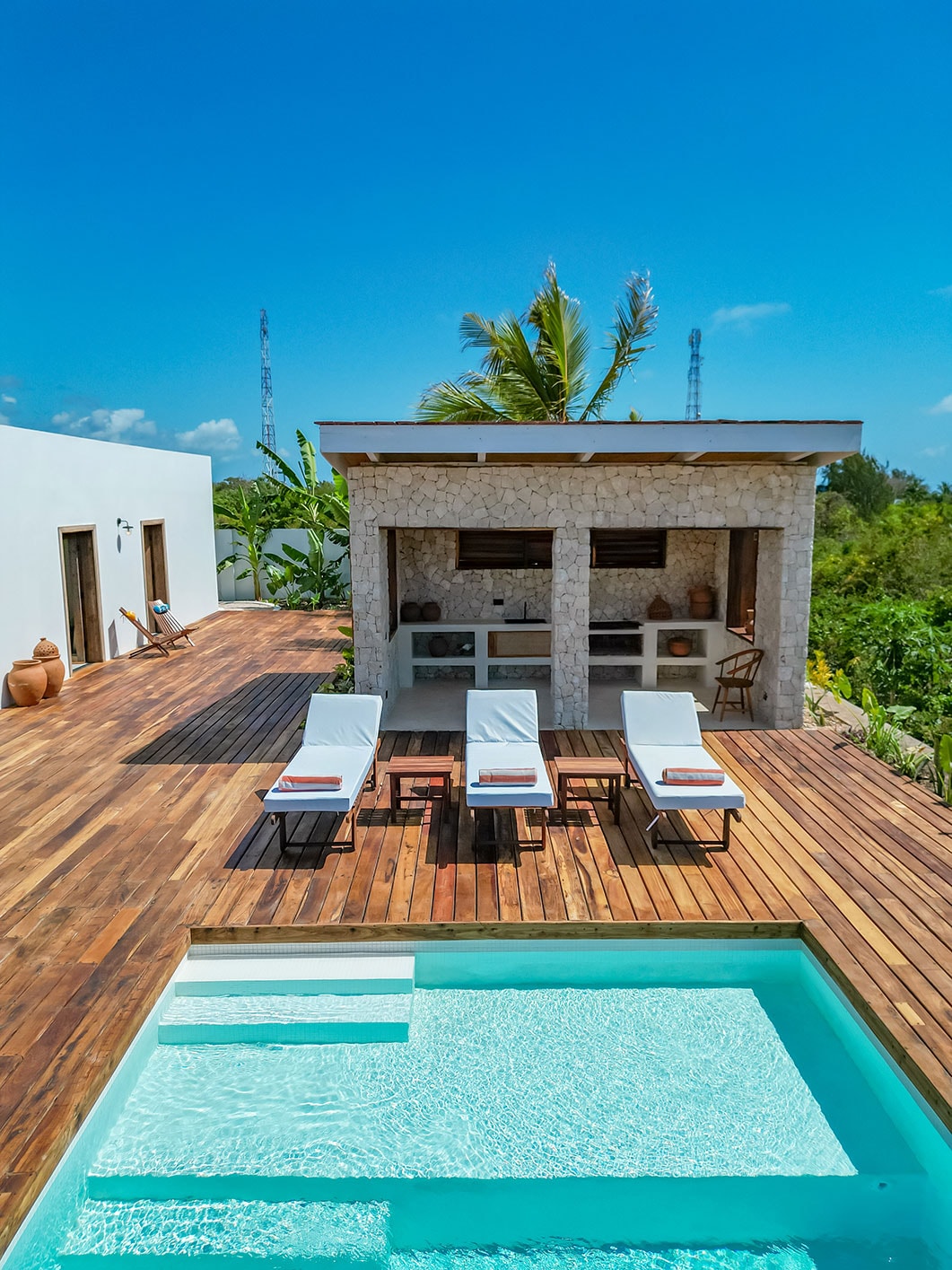 A serene outdoor area features a small swimming pool surrounded by wooden decking. Sun loungers with cushions are positioned nearby, while an outdoor kitchen area is visible in the background, complemented by lush greenery and a clear blue sky.