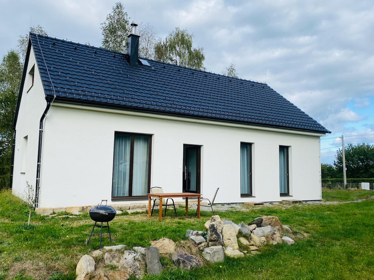 A modern, detached house with a simple design is set within a grassy area. Large windows allow for natural light, complemented by a small outdoor dining table and chairs. A fire pit is surrounded by rocks, enhancing the outdoor setting.