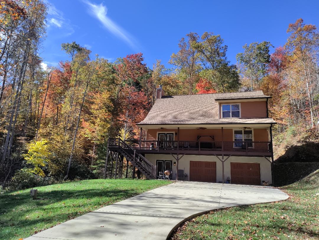 A two-story house is situated in a wooded area, surrounded by vibrant autumn foliage. The structure features a wraparound deck and a paved driveway leading to a garage. Sunlight casts a warm glow on the exterior, enhancing the inviting appearance of the home.