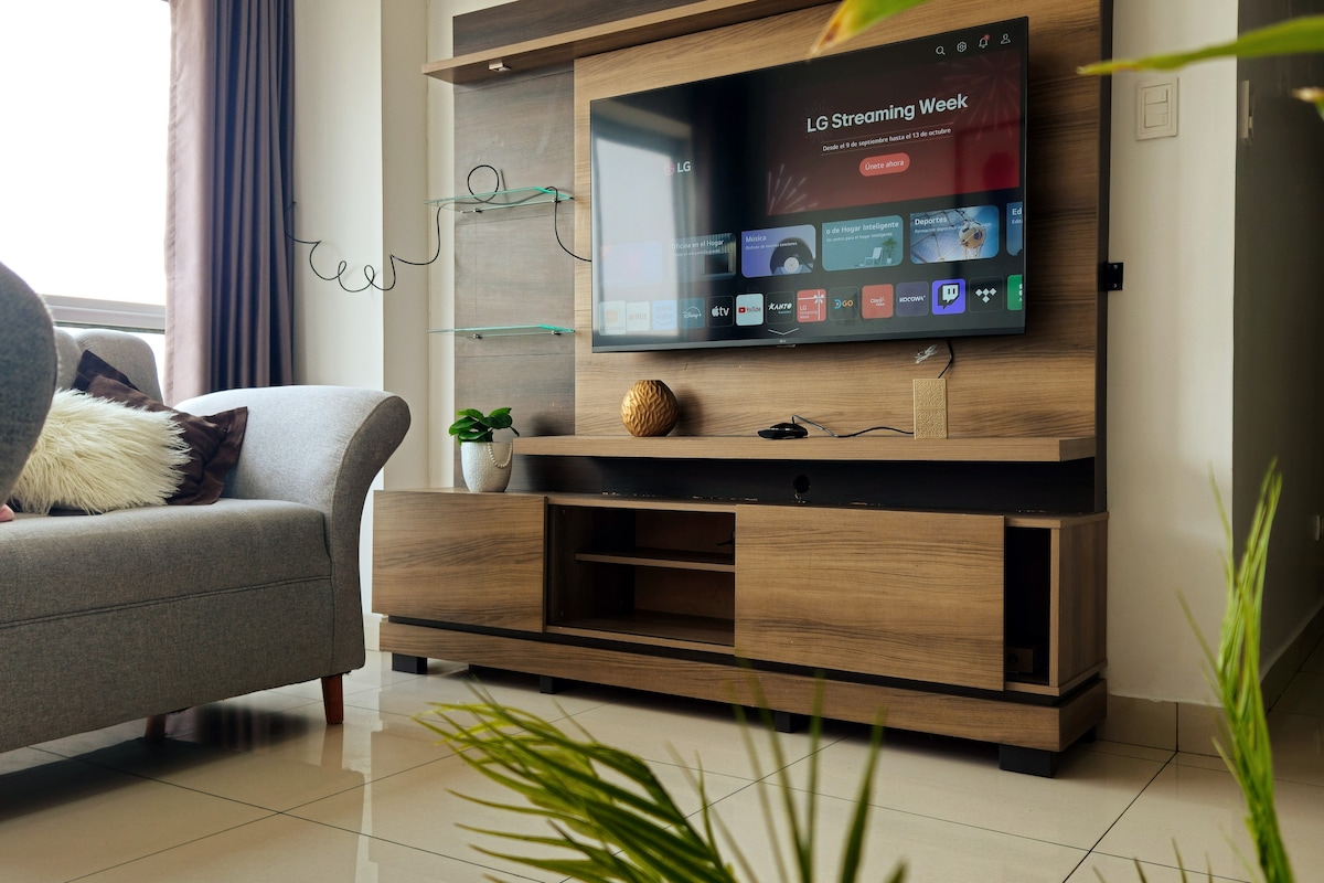 A modern living room is shown, featuring a sleek wooden entertainment center with a large flat-screen smart TV mounted above. A cozy gray sofa sits to the left, complemented by decorative cushions. Natural light filters through a nearby window with curtains, enhancing the space.