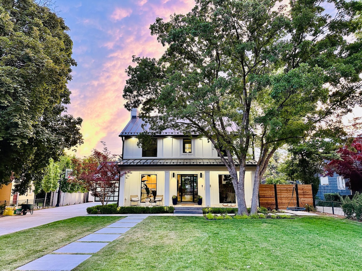 The exterior of the home features a modern design with large windows surrounded by lush greenery. A paved walkway leads to the entrance, framed by manicured lawns. The sky displays a blend of soft colors at sunset, enhancing the inviting atmosphere of the residence.