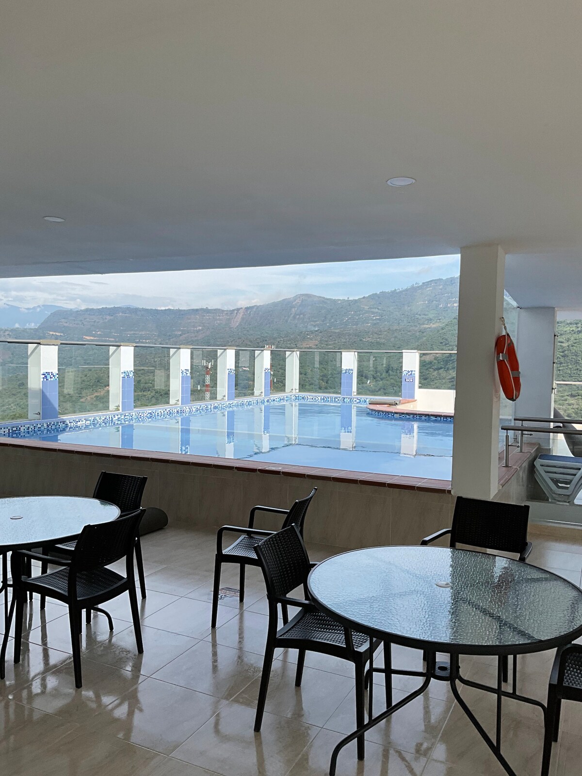 A spacious terrace area is visible, featuring several black chairs and round tables. A swimming pool with a blue mosaic border is seen in the background, bordered by glass panels that provide views of the surrounding mountains.