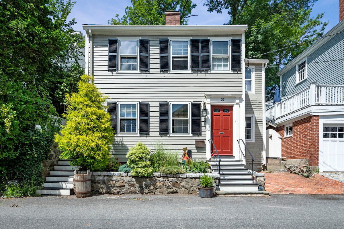 A charming colonial flat is presented, featuring a distinct gray exterior with black shutters. The vibrant red front door adds a welcoming touch. Lush greenery surrounds the entrance, highlighting the well-maintained landscaping and brick pathway leading to steps flanked by stone walls.