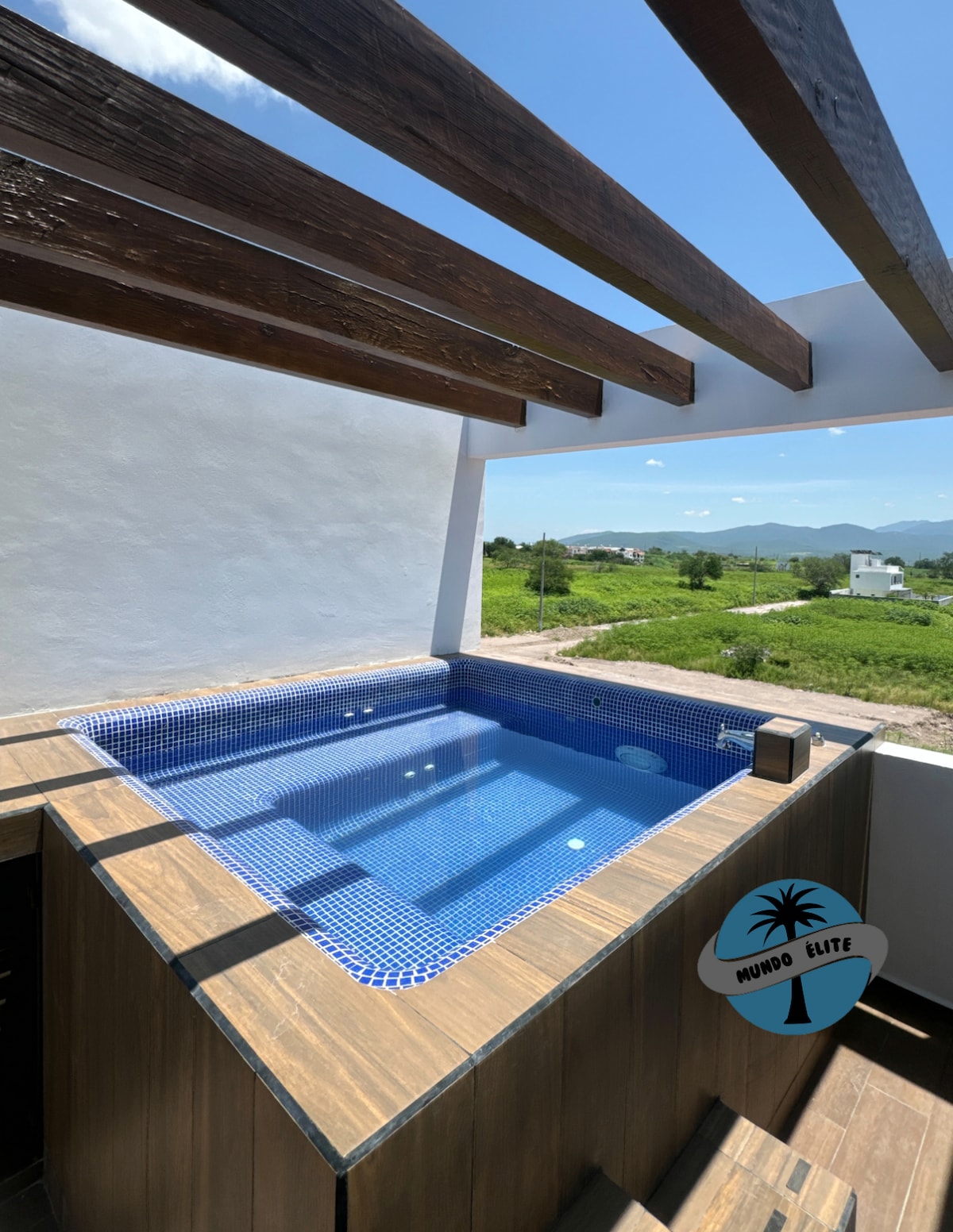 A private jacuzzi is situated under wooden beams, showcasing a deep blue tiled interior. Surrounding greenery is visible in the background, providing a serene natural setting. The space appears inviting and well-maintained, designed for relaxation during stays.