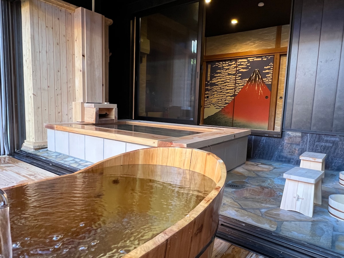 An indoor bathing area features a wooden hot spring tub filled with hot water, accompanied by a wooden washstand. A large glass wall allows natural light, while a striking mural depicting Mount Fuji adds cultural charm to the space.