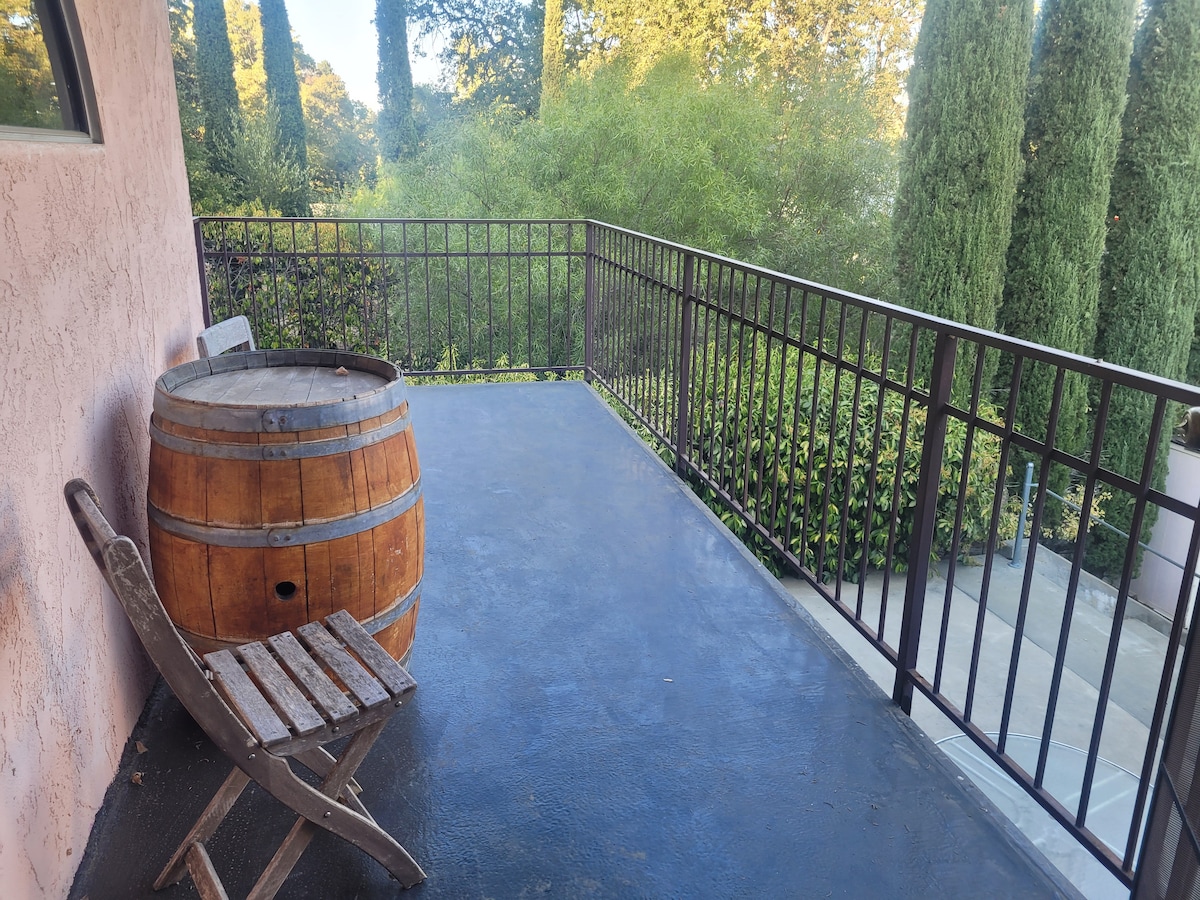 A private balcony area features a wooden barrel and a slatted chair, offering a space to enjoy views of the surrounding greenery. Tall trees can be seen in the background, contributing to a serene setting.