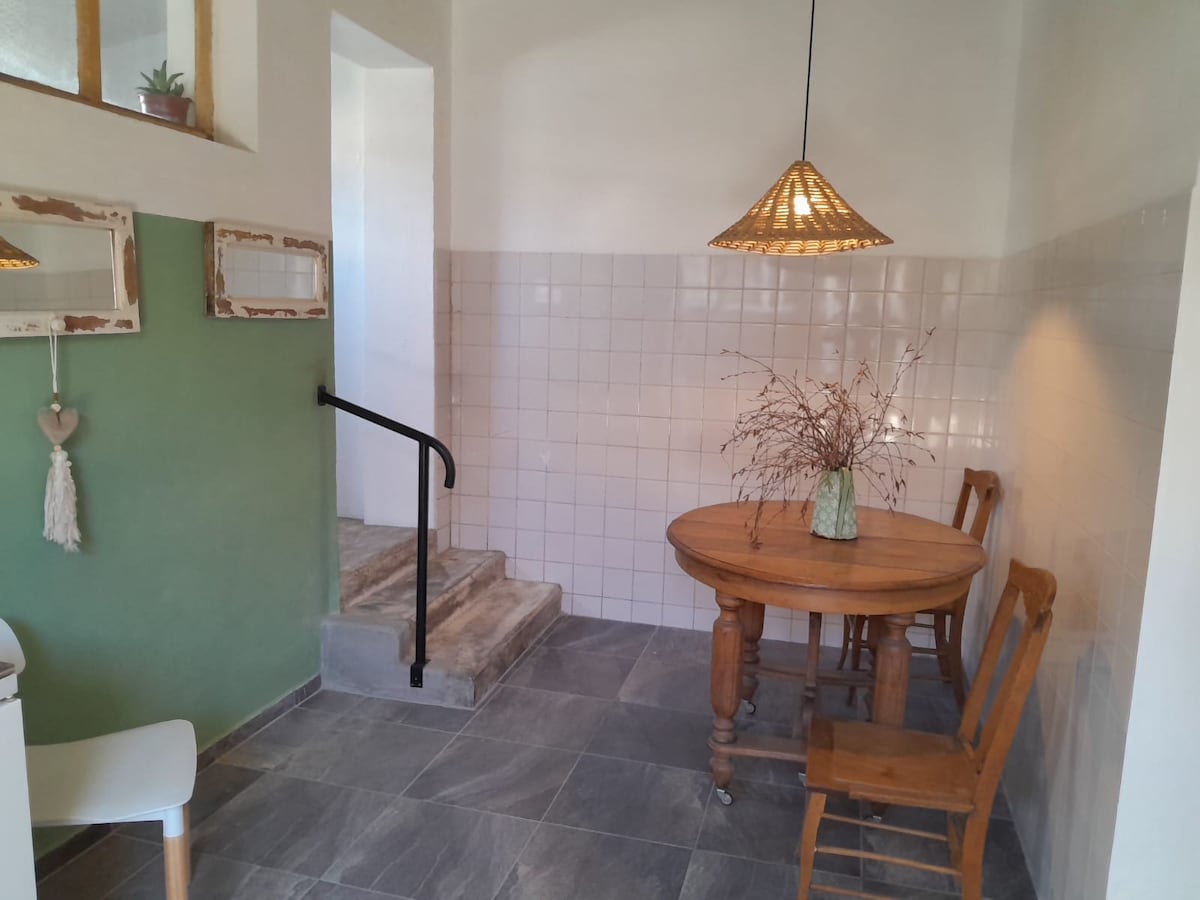 A bright dining area features a round wooden table surrounded by four wooden chairs. A decorative pendant lamp hangs above, and a small vase with dried flowers sits at the center. The walls exhibit a combination of white tiles and a muted green section, adding a touch of color.
