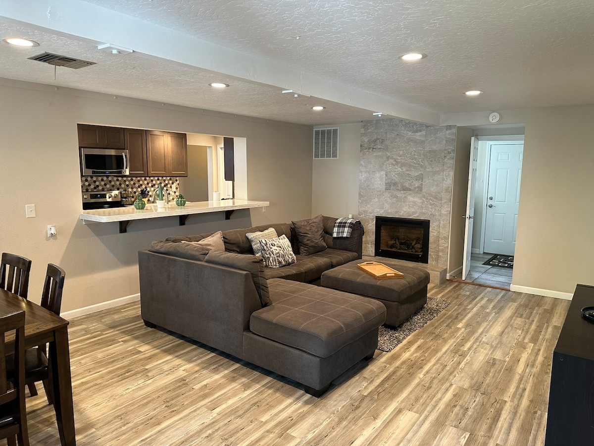 A cozy living area is presented with a large sectional sofa, complemented by a textured area rug. A stone fireplace serves as a focal point, while a bar counter transitions into a modern kitchen. Warm lighting illuminates the open space, inviting relaxation.