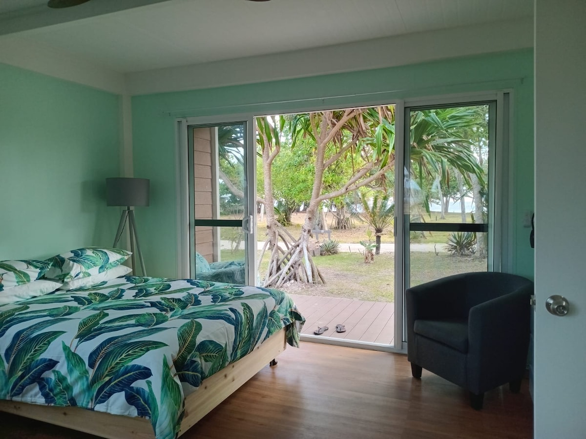 A light and airy bedroom features a spacious bed adorned with a leaf-patterned duvet cover. Large sliding glass doors provide a view of the outdoor area, where tropical greenery is visible. A cozy armchair complements the room's relaxed atmosphere.