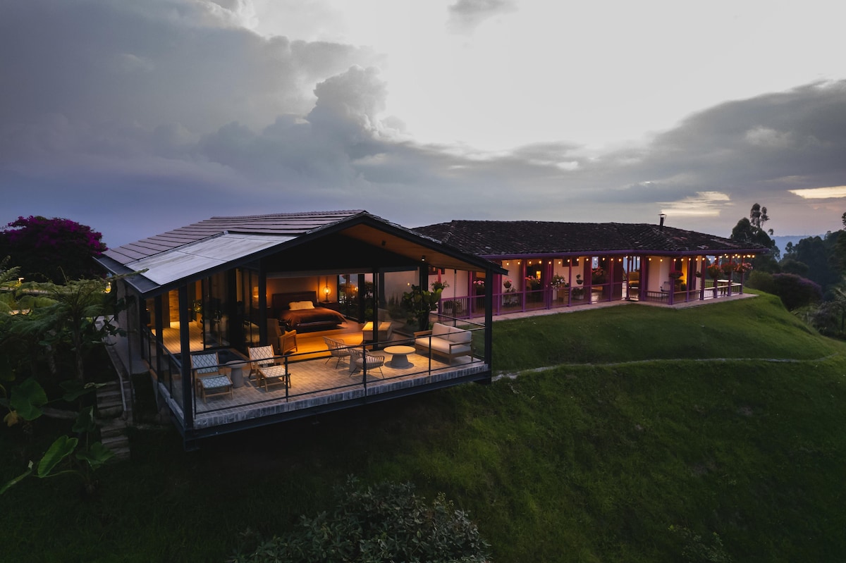 An aerial view of a modern house situated on a hillside. The structure displays large glass windows and a spacious outdoor terrace, illuminated during dusk. Surrounded by lush greenery, the exterior features a blend of contemporary design and natural elements.