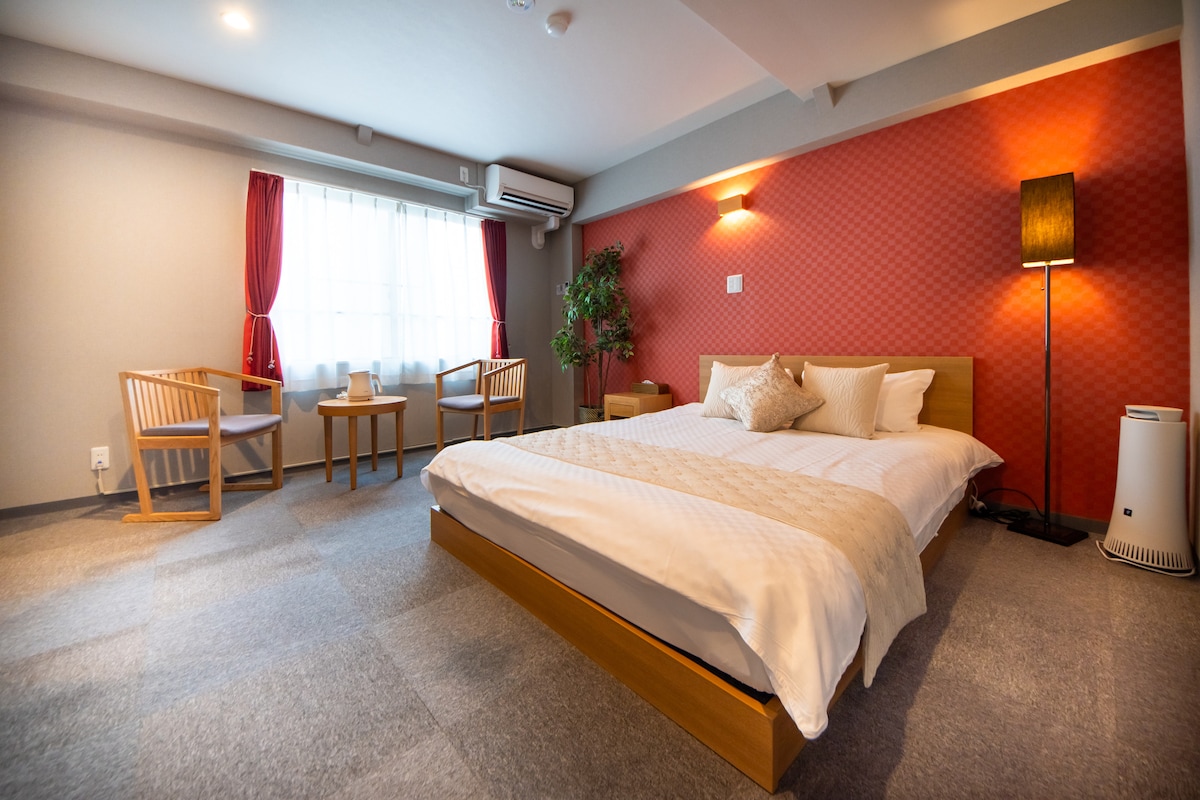 A spacious room is featured, showcasing a large bed with crisp bedding. Natural light filters through a window draped with sheer curtains. A cozy seating area includes two chairs and a small table, enhancing the room's comfort. A plant adds a touch of greenery.