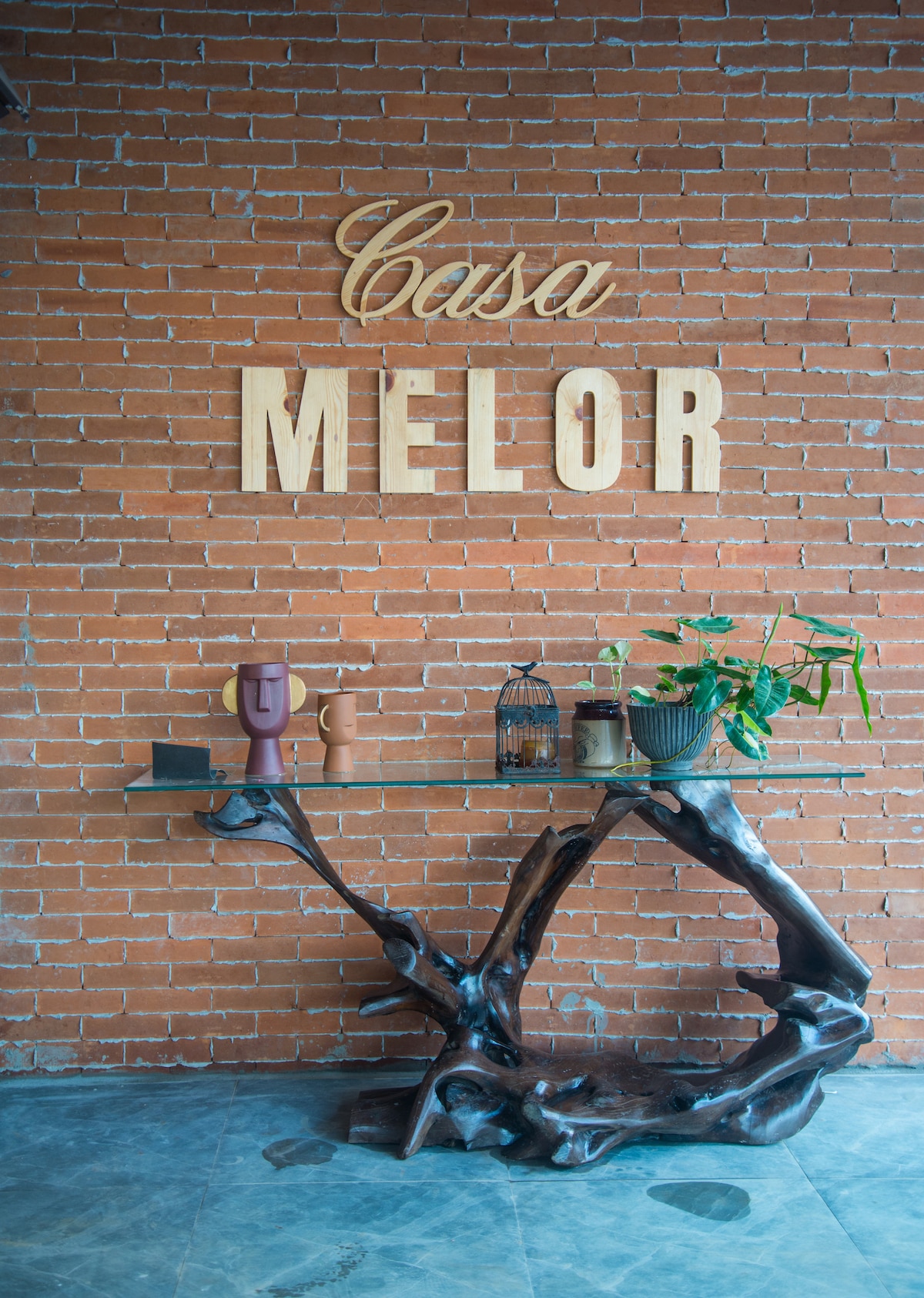 A welcoming entrance features the name 'Casa Melor' displayed prominently against a textured brick wall. A unique wooden console table with an organic shape supports decorative items, including ceramic vases and a small potted plant, creating a warm and inviting first impression.