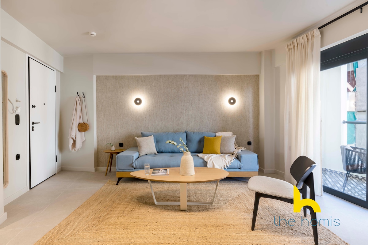 A cozy living room features a custom-made blue sofa paired with a light wood coffee table. Soft, stylish wallpaper surrounds the space, complemented by two wall-mounted lights. Natural light illuminates the area through sliding glass doors, leading to a small private balcony.