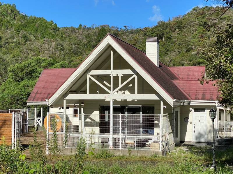 A spacious rental house is situated among greenery, featuring a distinct architectural style with a pitched roof and prominent front porch. Fencing surrounds the property, providing an enclosed outdoor area. Sunlight casts a warm glow across the exterior, enhancing the inviting ambiance of the home.