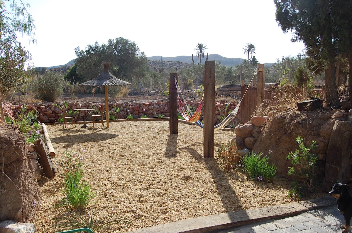 An outdoor space features a sandy area bordered by stone walls, with seating provided by a small wooden table and chairs. Two colorful hammocks are suspended between wooden posts. Native plants add greenery, and a thatched-roof gazebo is visible in the background.
