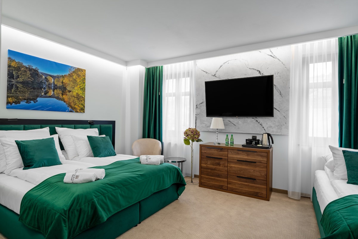 The room features two luxurious beds, one double and one single, dressed in white linens and complemented by green throws. A modern desk and a wooden dresser are present, alongside a large TV mounted on a marble wall. Natural light filters through sheer curtains.