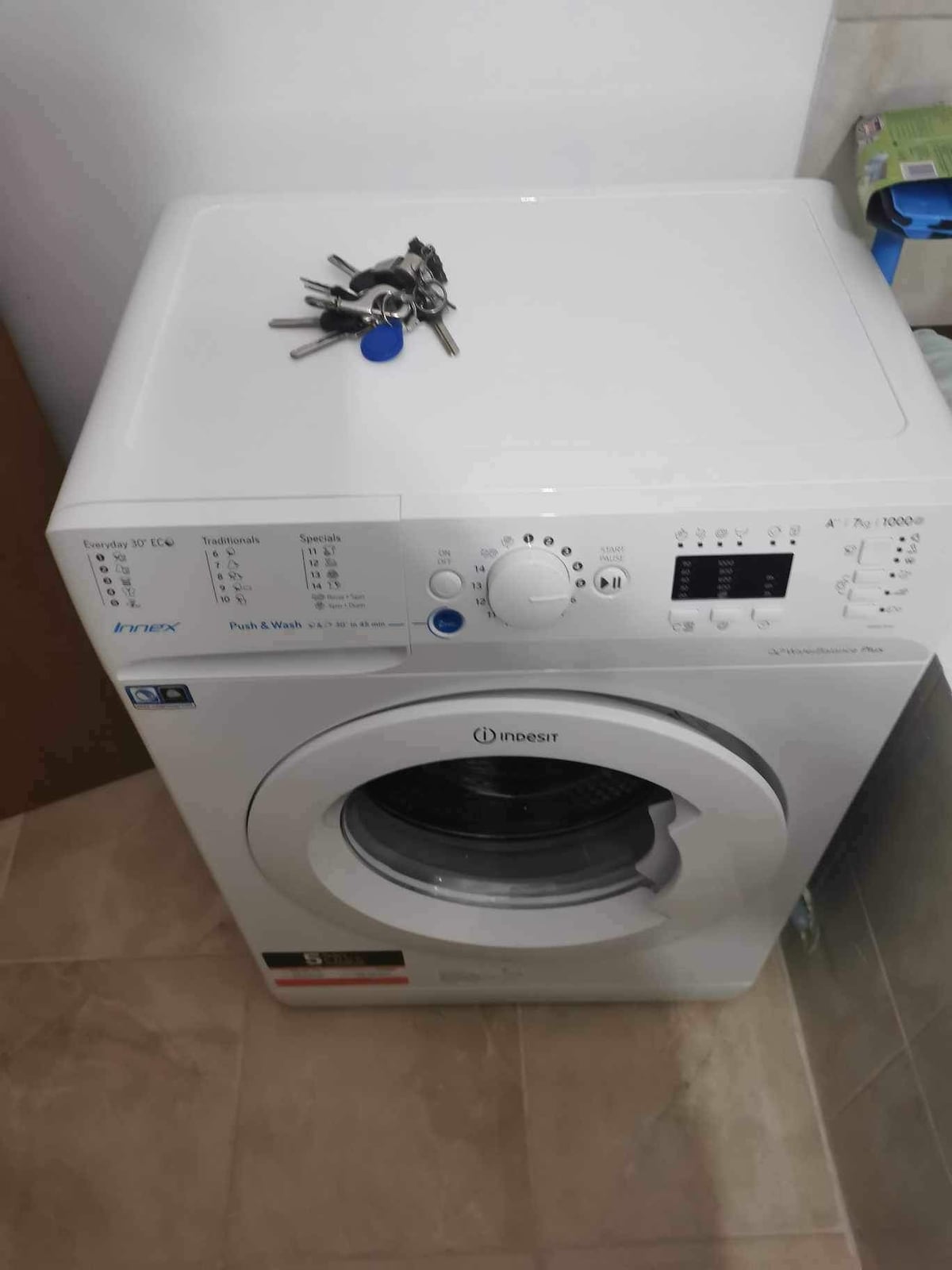 A compact washing machine is positioned against a wall, featuring a control panel with various settings and a round door showcasing the drum inside. A set of keys rests on top of the machine, highlighting the practicality of the space.