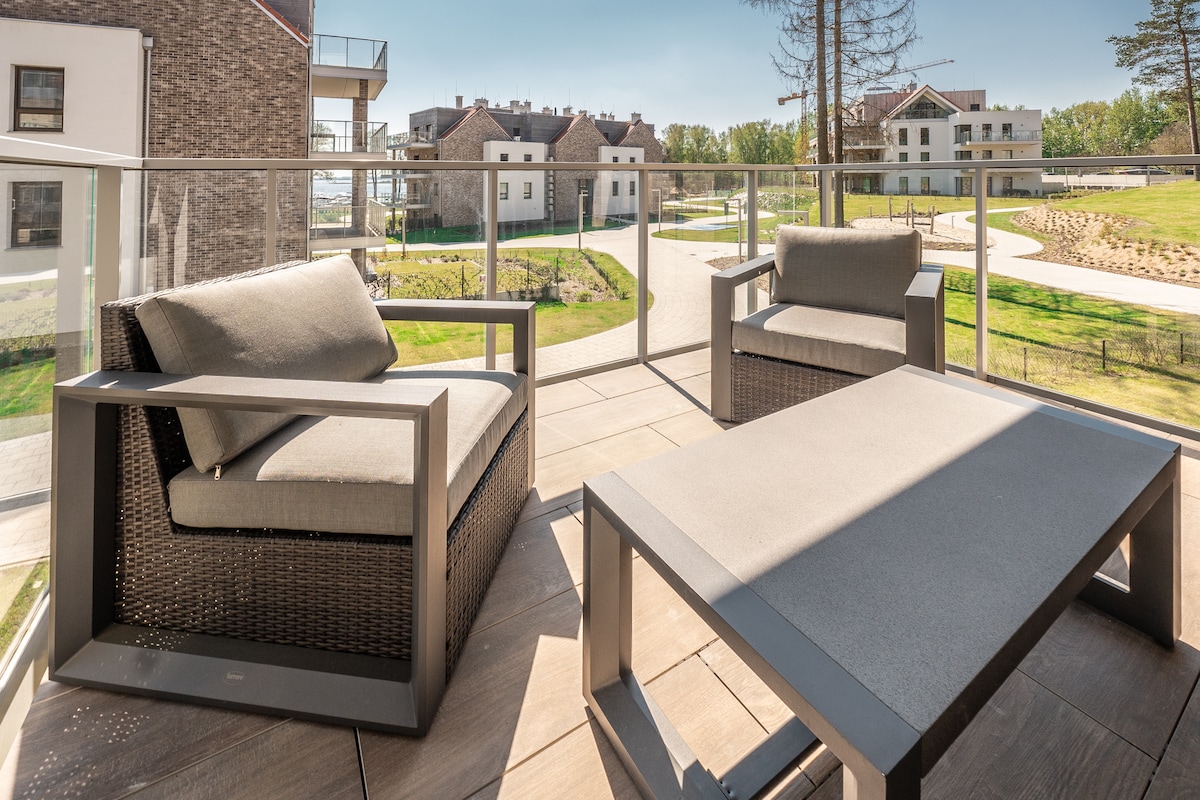 A terrace area is presented with modern outdoor furniture, including two comfortable armchairs and a low table. The setting offers clear views of the surrounding greenery and nearby buildings, creating an inviting space for relaxation.
