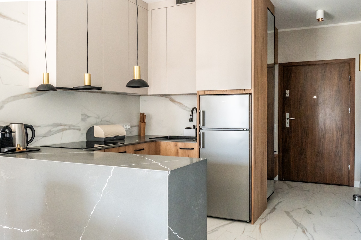 A modern kitchen featuring sleek cabinetry and stone countertops. Light fixtures hang above a spacious island, while essential appliances, including a refrigerator and coffee maker, are easily accessible. The kitchen is accented by a neutral color palette, creating a clean and organized cooking space.