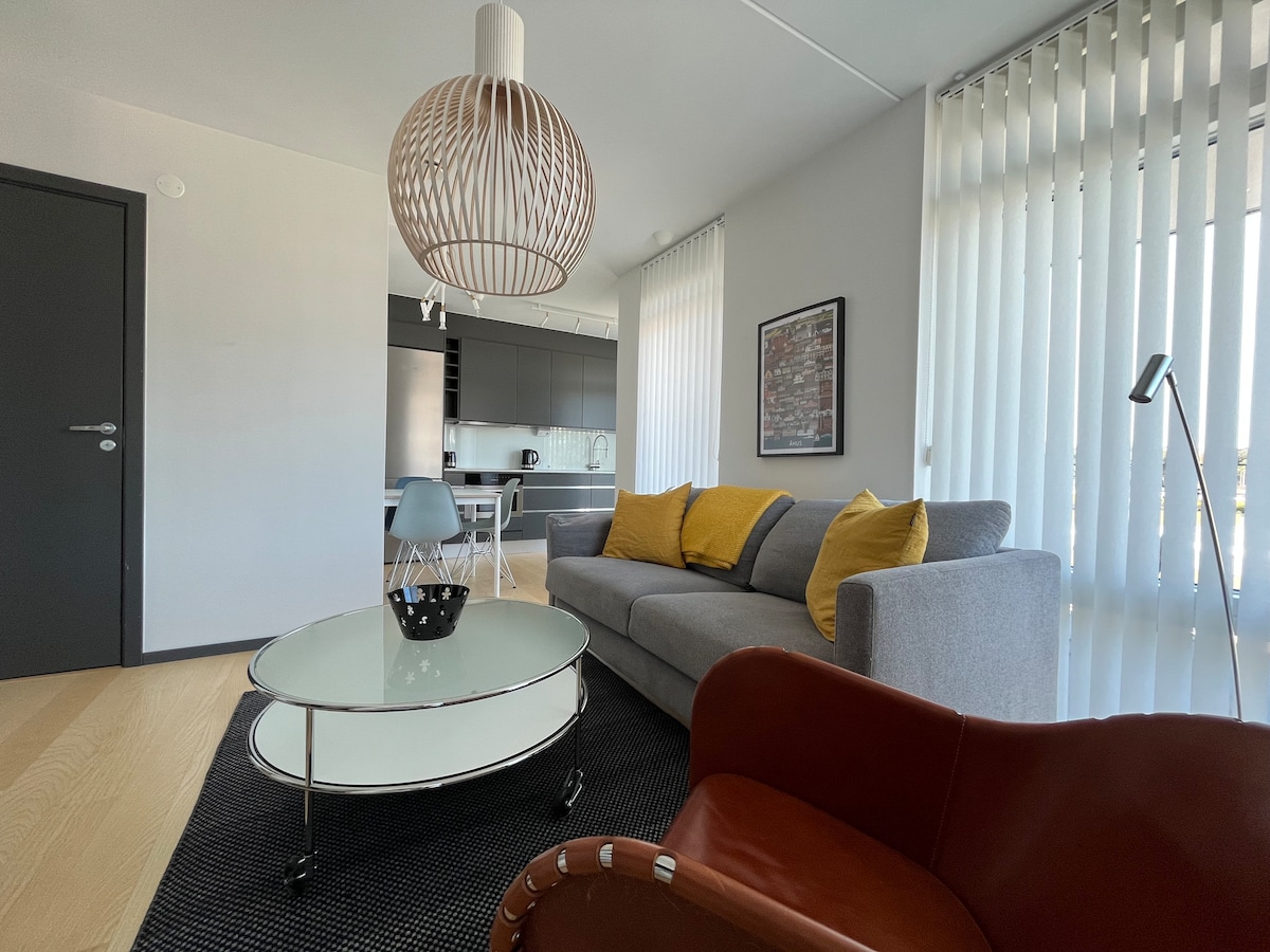 An inviting living area features a comfortable gray sofa adorned with yellow cushions. A modern coffee table rests on a textured rug, while a stylish pendant light hangs above. Light streams in through vertical blinds, illuminating the space. A glimpse of the kitchen can be seen in the background.