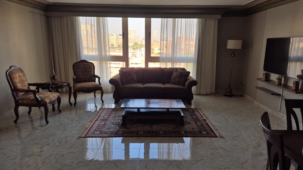 A spacious living area is highlighted by a comfortable brown sofa, two elegant armchairs, and a coffee table on a decorative rug. Large windows allow ample natural light and offer views of the city skyline. A wall-mounted smart TV is also visible.