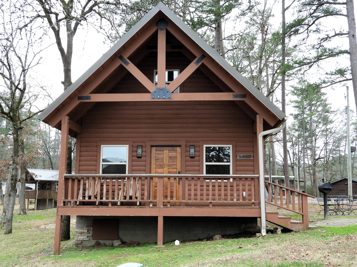 The exterior of a wooden cabin is showcased, featuring a prominent front porch supported by large beams and a symmetrical roof. Windows adorn each side, allowing natural light inside. A spacious yard is visible, surrounded by tall, leafy trees, enhancing the rustic setting.