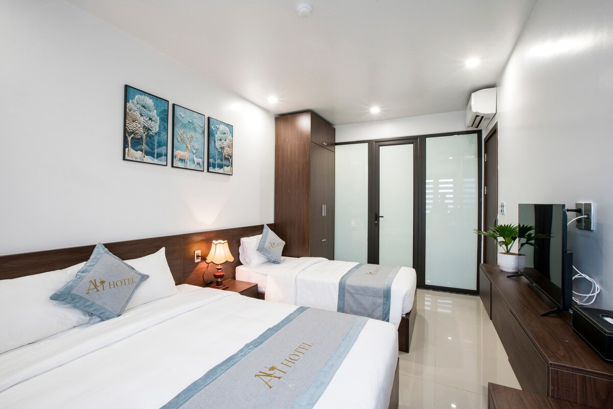 A well-appointed room is furnished with two twin beds topped with crisp linens and decorative pillows. A small bedside lamp provides soft lighting. The space features a wall-mounted television, a minimalist dresser, and a glass door leading to a separate area, complemented by artwork on the walls.