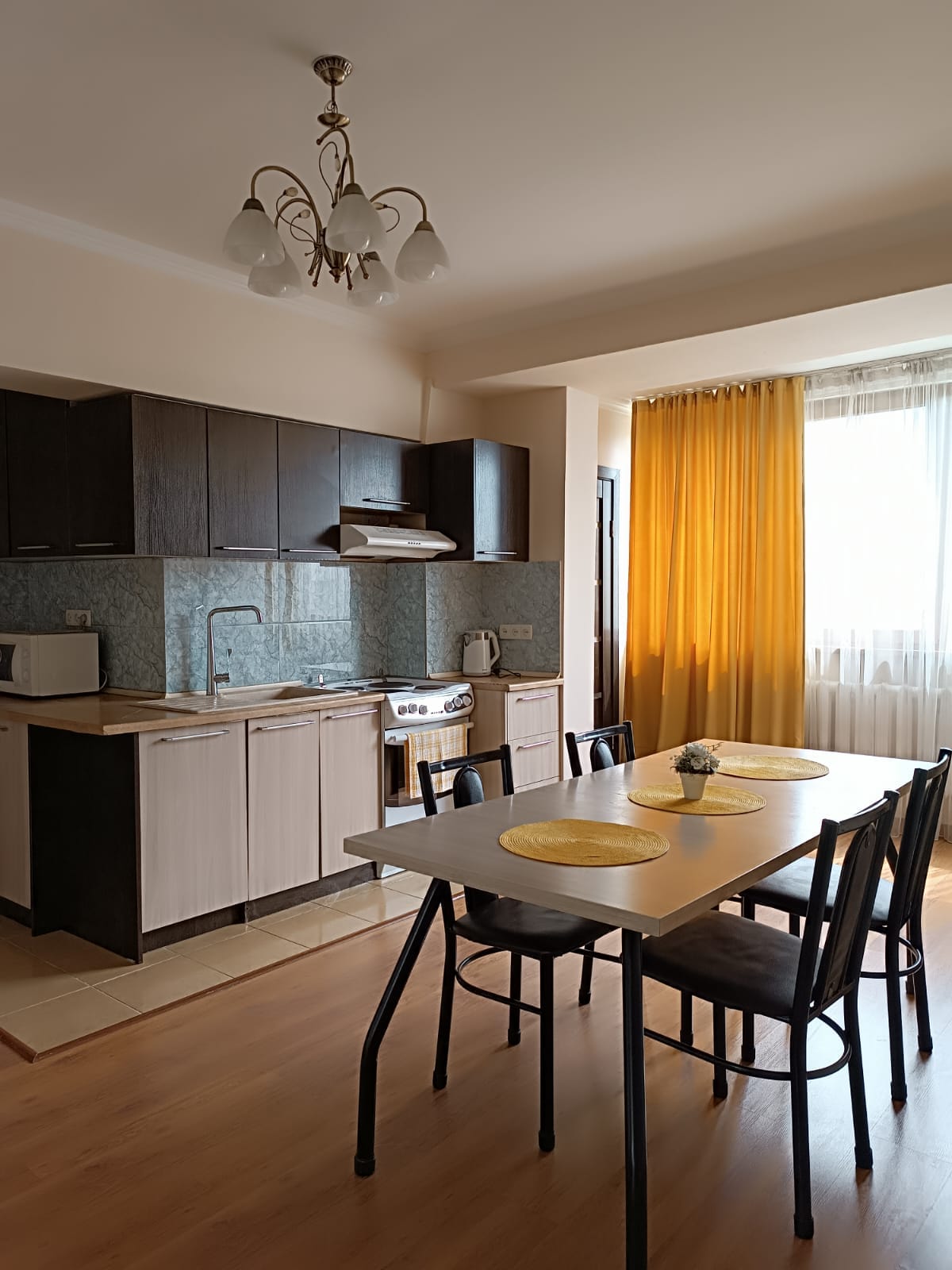 A modern kitchen area features dark cabinetry and a light-colored countertop. A dining table with four black chairs is set with yellow placemats. Large windows allow natural light to enter, complemented by sheer curtains and vibrant yellow drapes.